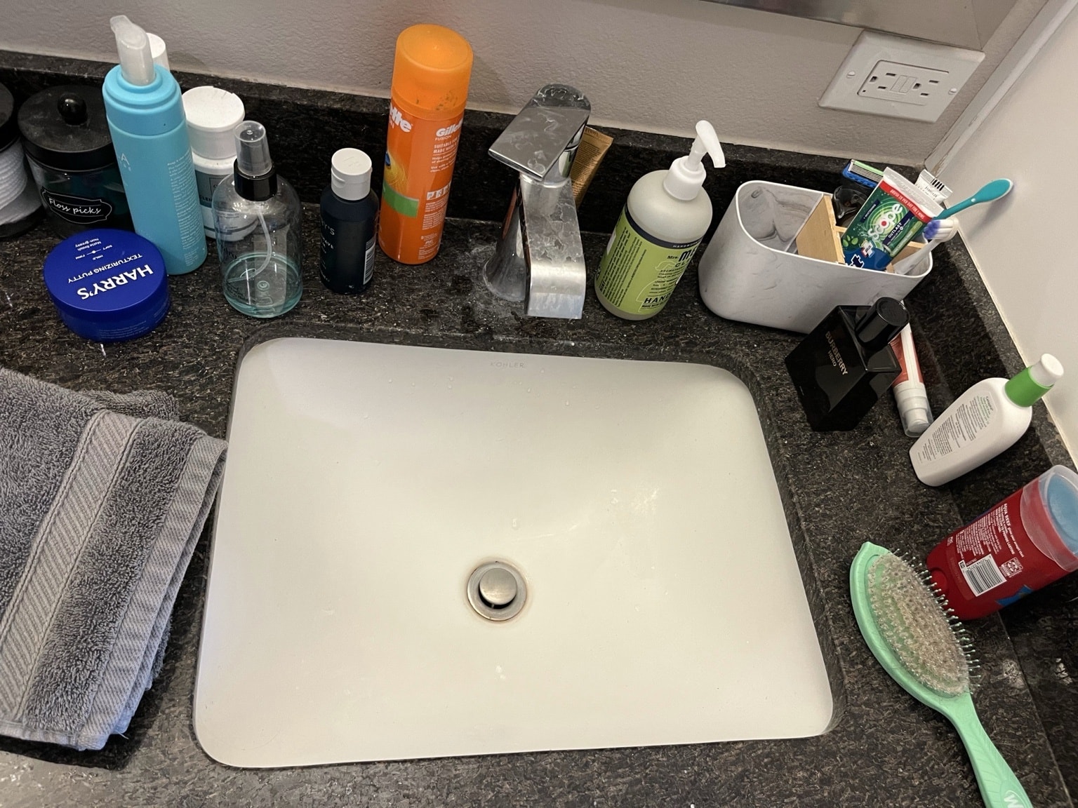 Bathroom sink before cleaning — cluttered dark granite counter with grooming products